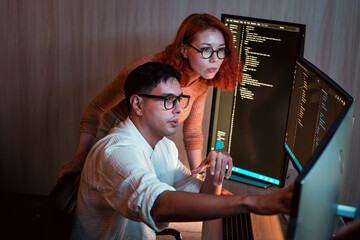 Two software developers collaborate at dual monitors in modern office at night. Coding, debugging, and programming show teamwork, technology, and startup energy in an intense workspace setting.