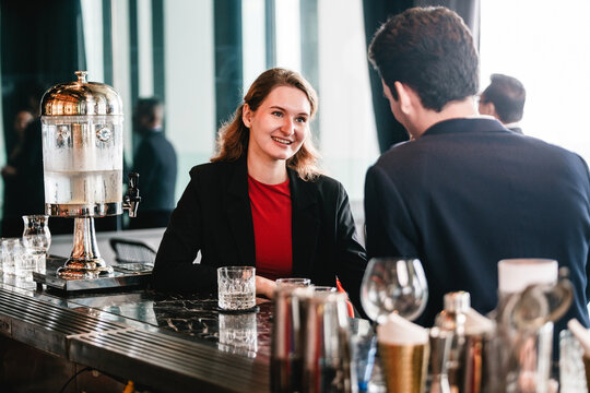 Two business colleagues network at modern hotel bar, seated across counter with glassware. Informal meeting blends corporate partnership, hospitality, and professional teamwork discussions.