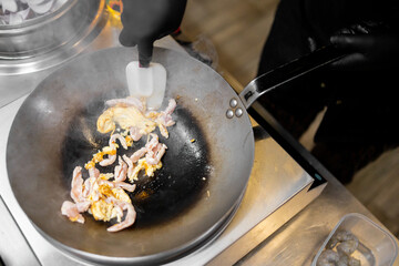 Chef wearing black gloves cooks chicken in a wok on stovetop, with steam rising. Fresh ingredients like onions and shrimp visible. Professional kitchen scene, close-up