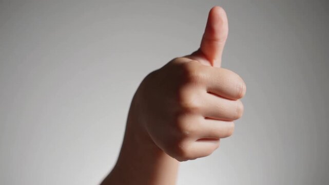 The hand of juvenile or child's hand displaying affirmative thumbs-up gesture on white and gray backdrop