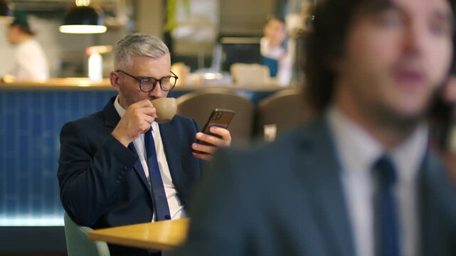 Middle aged businessman in suit and glasses drinking coffee and browsing the web on his smartphone while sitting at table in modern cafe