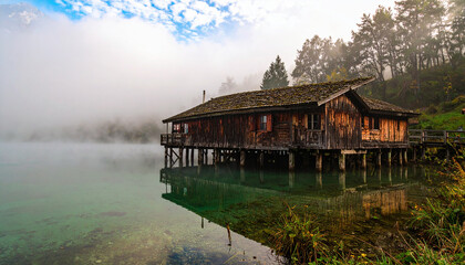 Isolated wooden house on stilts in misty water, surrounded by tranquil nature, lush green trees, and calm reflection, creating peaceful cinematic atmosphere