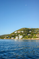 Fototapeta premium Picturesque hillside neighborhood with modern houses overlooking the ocean in Knysna. Clear blue sky with the moon above, lush greenery, and sparkling water create a serene coastal landscape