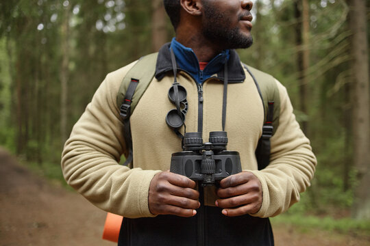 Black young adult man standing in forest holding binoculars with both hands, wearing backpack and outdoor gear, looking away from camera, exploring nature during hike - Powered by Adobe