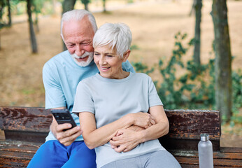 Happy active senior couple having fun using smartphone and wearing sportswear, after having an exercise sport activity outdoors