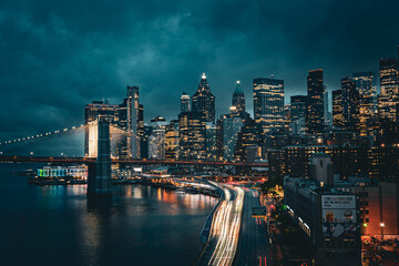 New York City skyline with Brooklyn Bridge at night, light trails from traffic below.