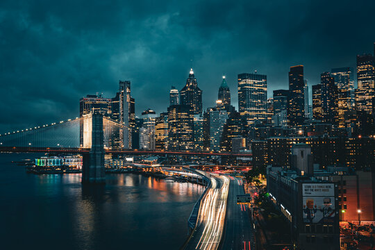 New York City skyline with Brooklyn Bridge at night, light trails from traffic below. - Powered by Adobe