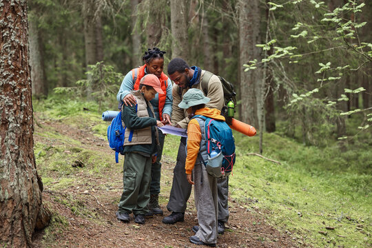 Black man and Black woman guiding two Black children while studying map during forest hike, all wearing backpacks and outdoor gear, standing together on woodland trail