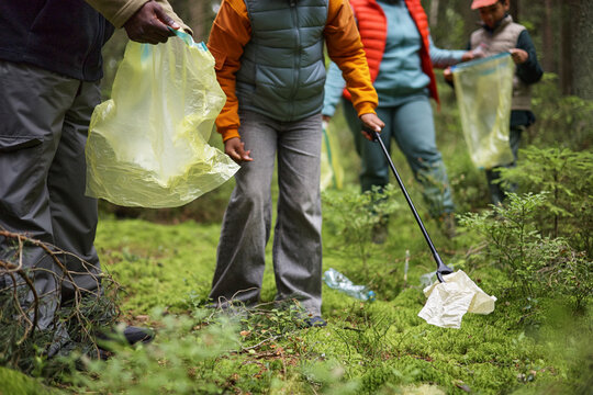 Unrecognizable family members cleaning forest floor, collecting litter with plastic bags and grabbers, focusing on teamwork and environmental conservation outdoors