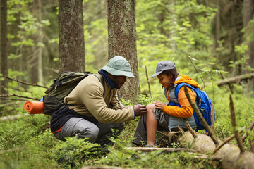 Black middle aged man kneeling in forest helping Black child with injured knee during hiking trip,...