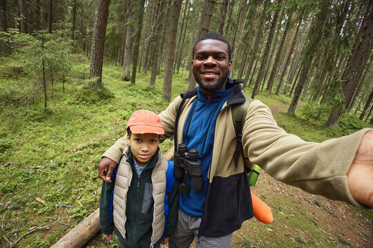Black man and boy standing together in forest, man smiling and taking selfie while boy looking at camera, both wearing outdoor clothing and carrying hiking gear