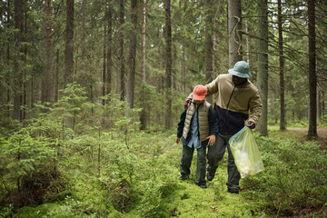 Obraz premium Black man walking with child through forest, holding plastic bag and picking up litter, both wearing hats and outdoor clothing, surrounded by tall trees and green vegetation