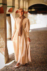 Smiling Muslim woman in modest beige dress and hijab standing on cobblestone floor under traditional arches, cultural fashion and heritage portrait.