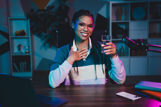 Young female podcaster with microphone and glass of water recording at her desk in a stylish neon-lit studio