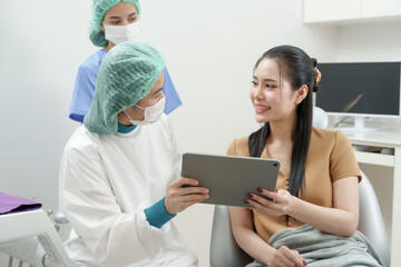 Asian dentist wearing surgical gown and mask showing digital x-ray to female patient using tablet in dental chair, discussing treatment plan with nurse assistant standing nearby in dental clinic