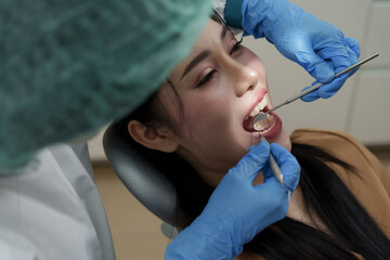 Asian female patient lying on dental chair receiving oral examination by dental assistant wearing surgical cap and blue gloves. Assistant using dental mirror and scaler to check teeth and gums