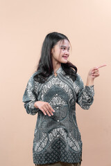 An Asian woman wearing a patterned batik shirt points to the side with a friendly smile, showing elegance, confidence, and positive body language in a modern professional concept.