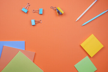 Top view of vibrant office supplies on orange background: colorful paper clips, pens, sticky notes, and stacked paper sheets. Ideal for business organization, planning, and stationery concepts