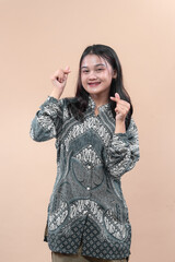 Fototapeta premium Smiling young Asian woman wearing elegant batik shirt making finger heart gesture with both hands, showing happiness, confidence, and cultural pride suitable for global advertising concepts.