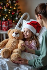 A little girl wearing a red Santa hat lies in a hospital bed, smiling as a nurse gives her a big teddy bear. The background shows a decorated Christmas tree and wrapped presents