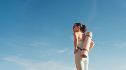Sporty woman carrying a yoga mat, blue sky background