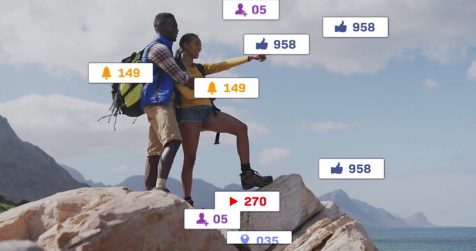 Pointing couple standing atop cliff overlooking ocean, with hiking backpacks, social media icons