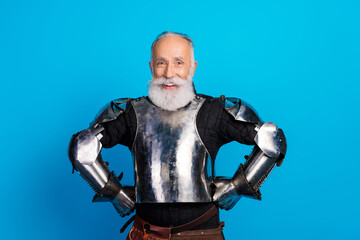 Obraz premium Smiling elderly man dressed as medieval knight in armor on a vibrant blue background showcasing costume and historical reenactment
