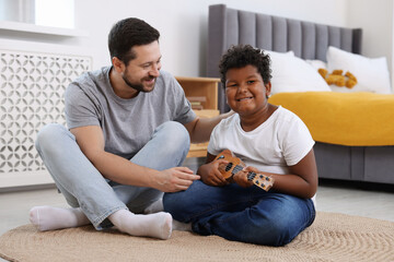 Adoption. Father teaching his son to play ukulele on floor at home