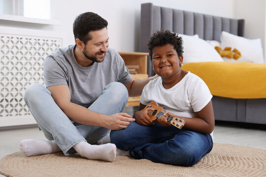 Adoption. Father teaching his son to play ukulele on floor at home