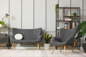 Stylish grey armchair, sofa, shelving unit and houseplants indoors. Interior design
