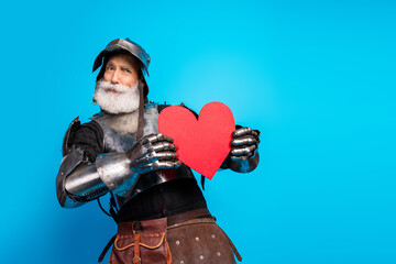 Senior knight in armor holding a red heart on a blue background symbolizing bravery, love, and chivalry
