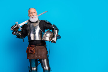 Obraz premium Portrait of a senior man in full medieval knight armor holding a sword and helmet with bright blue background