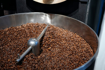 Dark roasted coffee beans swirl in a large metal roaster, releasing rich aromas. The process occurs in a lively cafe, filling the air with an inviting scent in the morning hours