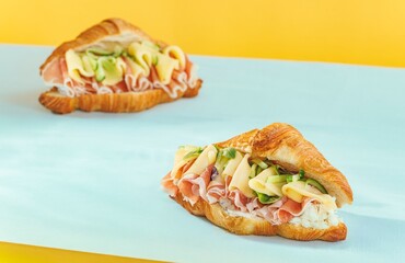 Two golden croissants, bursting with savory fillings like cheese slices, thin-shaved ham, and ribbons of fresh zucchini, rest on a contrasting blue and yellow surface