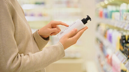 Close-up of female customer holding a cosmetic bottle and reading the label. Woman choosing shampoo, facial cleanser, body wash or conditioner in a cosmetics store. Conscious choice and personal care.