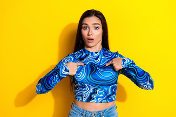 Surprised young woman in blue patterned top making expressive gestures against vibrant yellow background