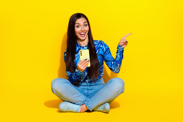 Attractive woman sitting cross-legged on yellow background holding smartphone in hand and pointing energetically to side.