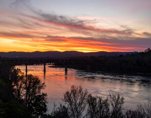Obraz premium Żywe kolory zachodzącego słońca malują niebo nad rzeką i wiaduktem kolejowym