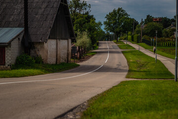 A peaceful countryside scene featuring a winding asphalt road, an old barn, green lawns, and a solitary pedestrian under cloudy skies.