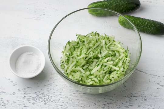 Grated cucumber in glass bowl with salt on white table for salad or tzatziki preparation