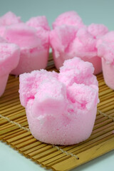 A close-up of a vibrant pink Kue Mangkok or Huat Kueh, a traditional steamed sponge cake from Southeast Asia with a signature cracked top. The cakes are on a bamboo mat with a plain background.
