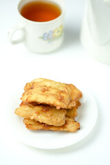 A small stack of fried banana fritters on a white plate, with a blurry cup of tea and a teapot in the background.