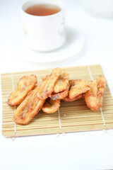 A pile of fried banana fritters on a bamboo mat with a blurry white teacup and saucer in the background.