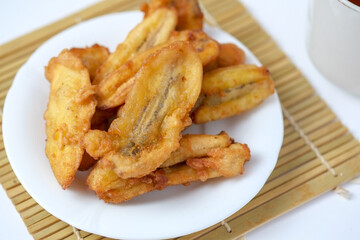 A white plate holds a pile of golden-brown fried banana slices, sitting on a bamboo mat.
