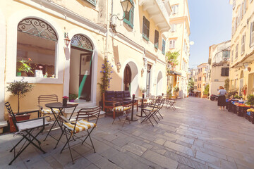 Inviting outdoor cafe terrace with wrought iron bistro tables and striped chairs along cobblestone pedestrian street in historic Corfu old town.