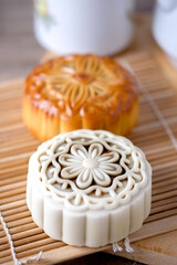 wo elegant mooncakes, one snow skin and one baked, sit on a bamboo mat with a tea set in the background, capturing a cozy mid-autumn festival moment.