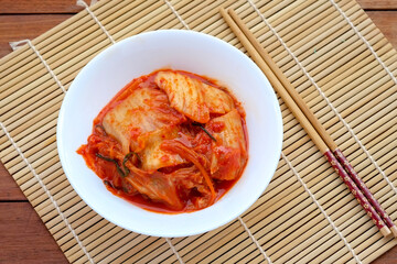 A white bowl filled with spicy red kimchi sits on a woven bamboo mat next to a pair of chopsticks...
