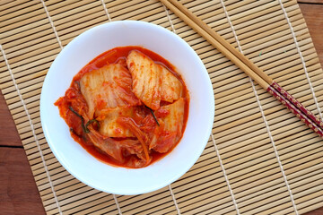 A white bowl filled with spicy red kimchi sits on a woven bamboo mat next to a pair of chopsticks...