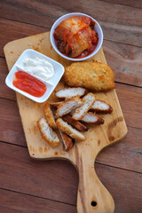 Sliced home made  fried chicken cutlet, with bowls of kimchi and two sauces, are served on a wooden cutting board.