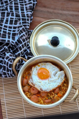 A bowl of instant korean ramen or noodles topped with a fried egg and sliced sausage, served in a brass pot with a lid on a bamboo mat.